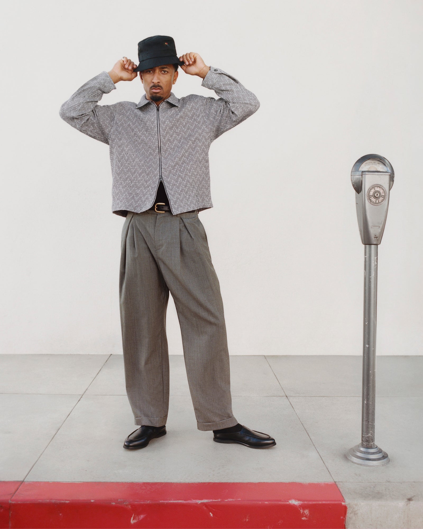 Man in Textured Zip Shirt, wide trousers, and black hat stands by a parking meter on a sidewalk with a red curb.