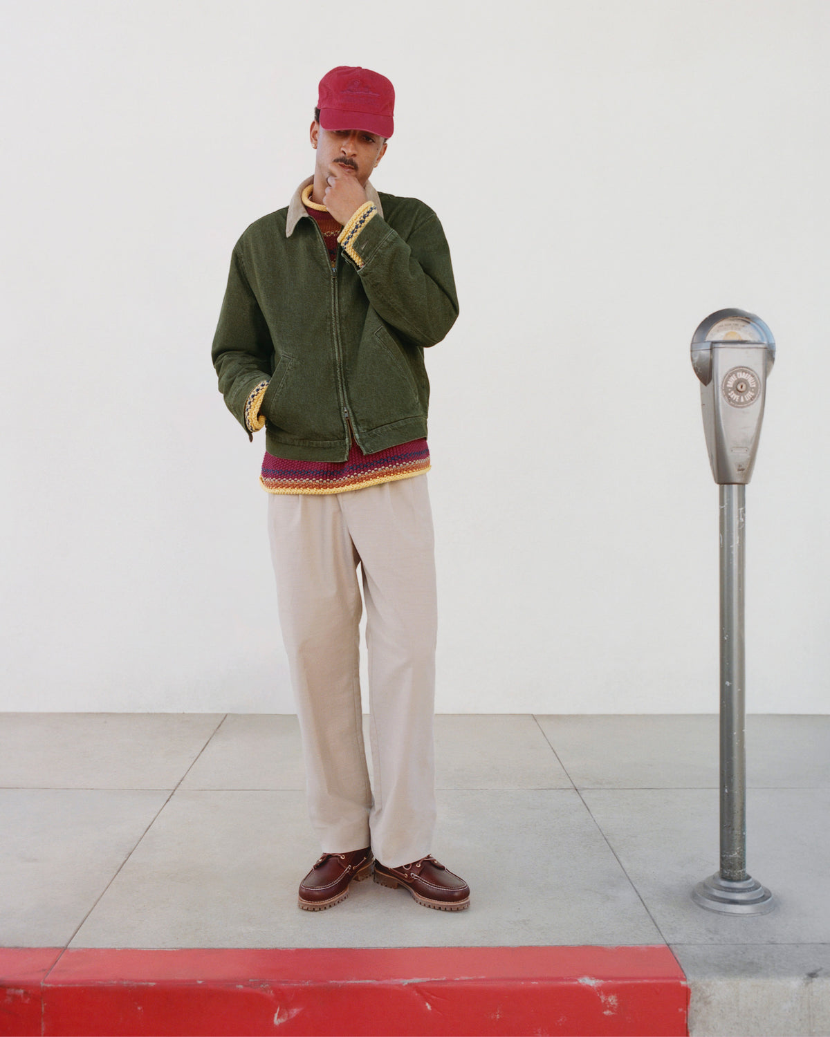 A person in a Sun Faded Unisphere Hat and green jacket stands by a parking meter on a sidewalk with a red curb.