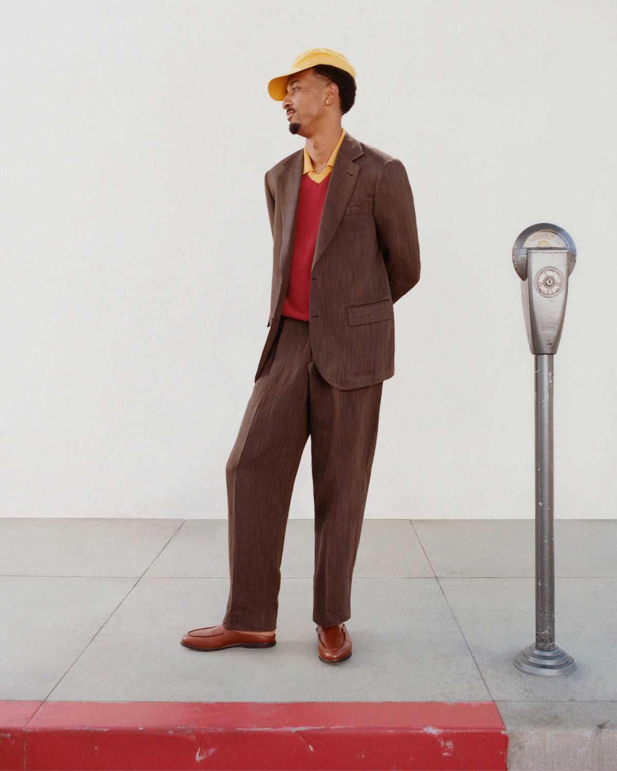 A man in a Knit Sport Pullover and yellow cap stands on a sidewalk by a parking meter, facing away.