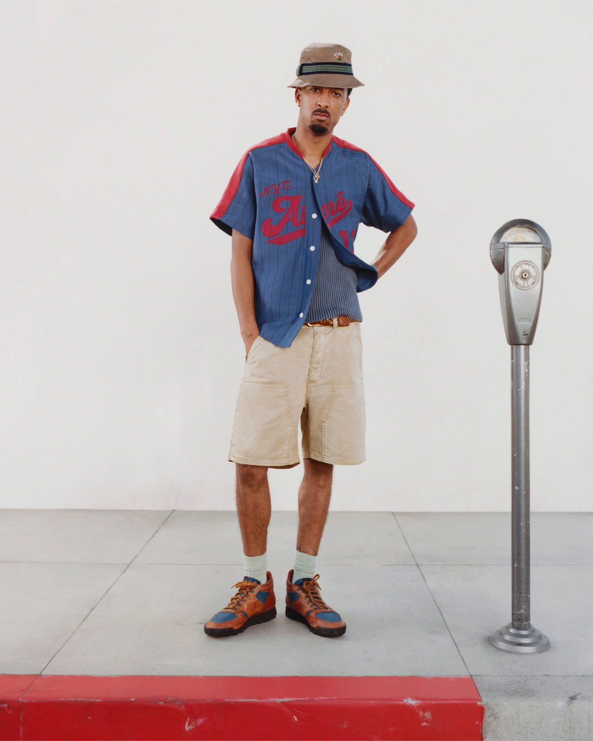 A man in a blue shirt and Garment Dyed Carpenter Short stands by a parking meter on a sidewalk with a white wall behind.