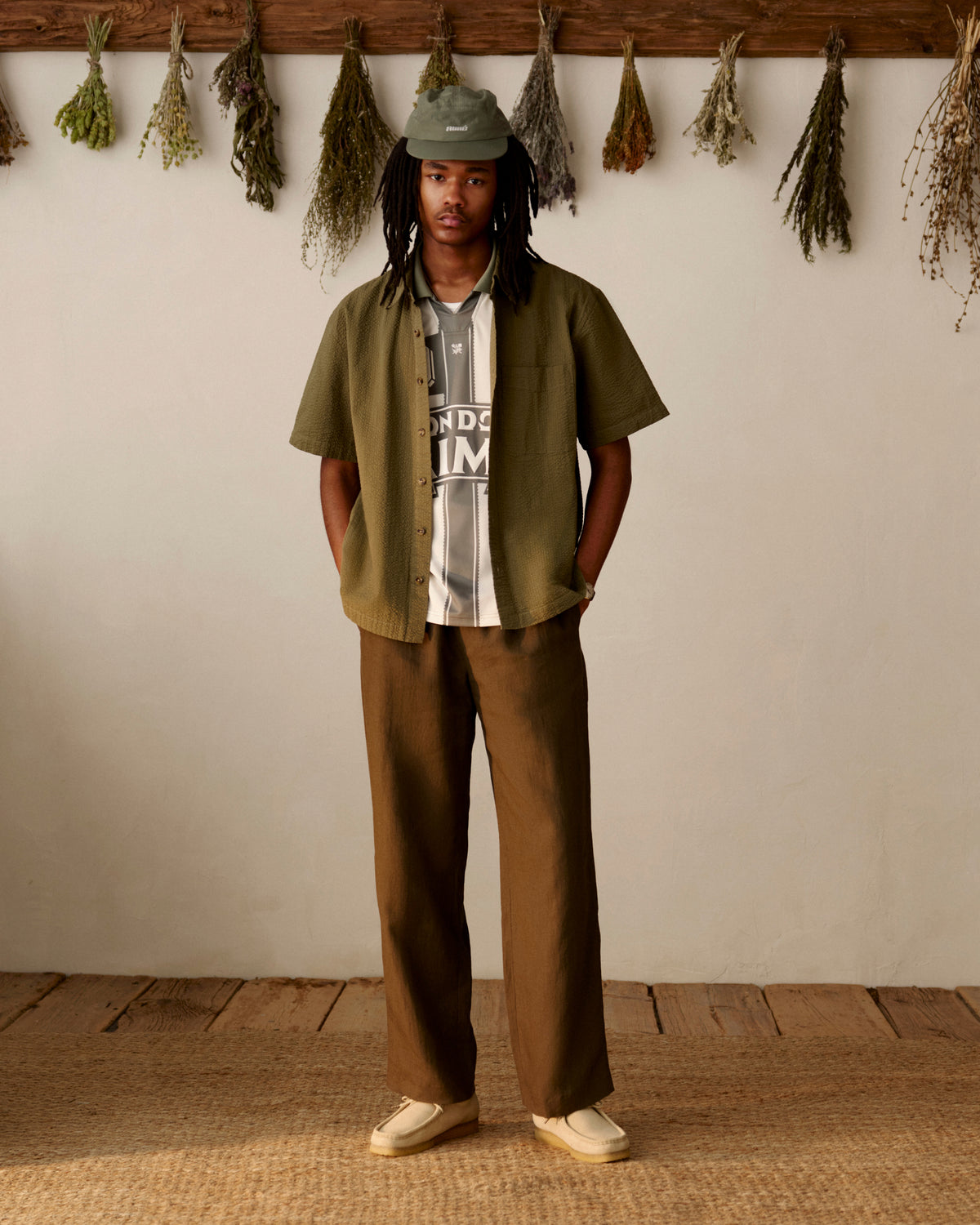Person in green hat, Short-Sleeve Seersucker Shirt, brown pants, and white shoes stands indoors under dried plants.