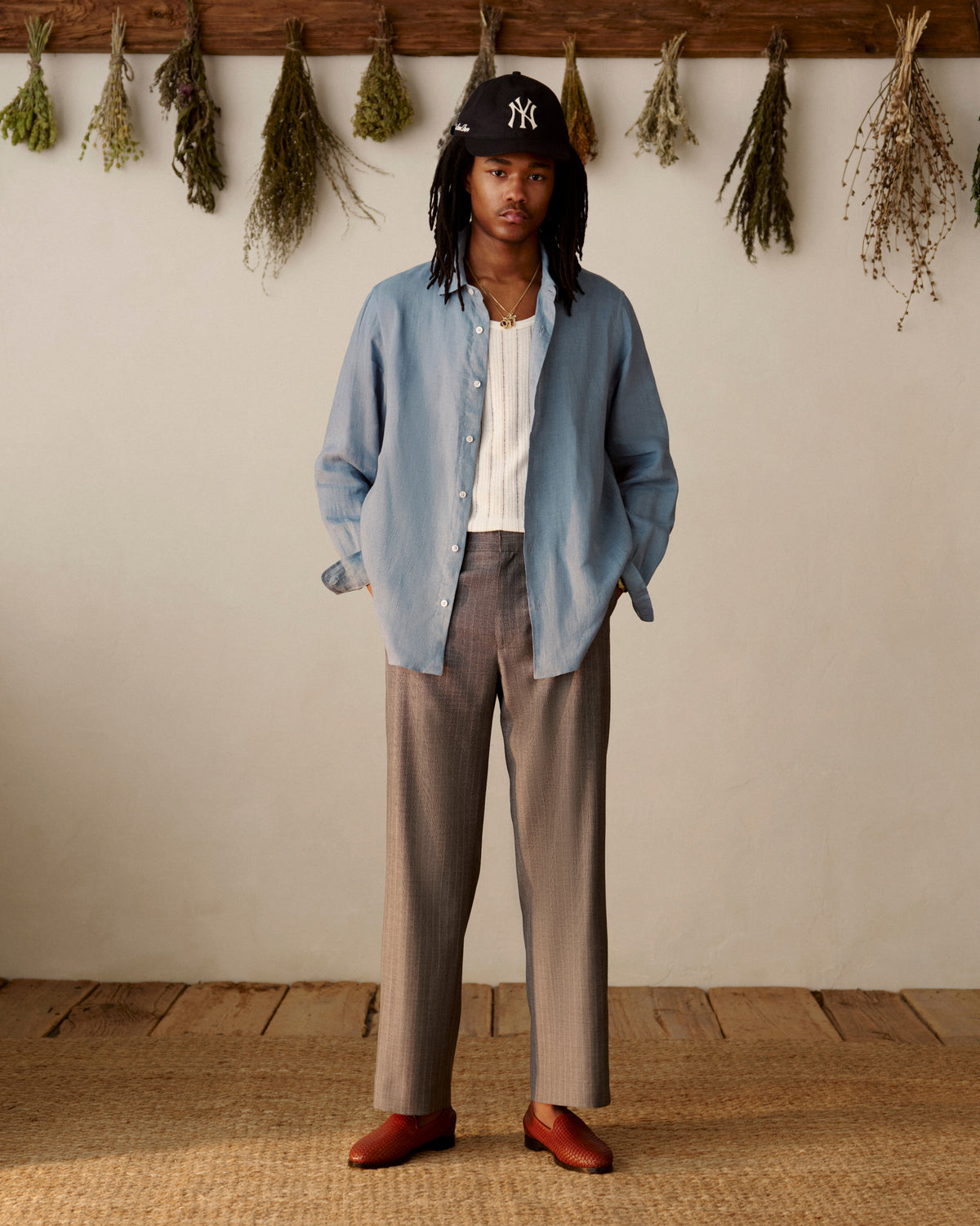 A person wears a blue shirt, gray pants, Yankees cap, and Leather Raffia Loafer with dried herbs hanging behind them.