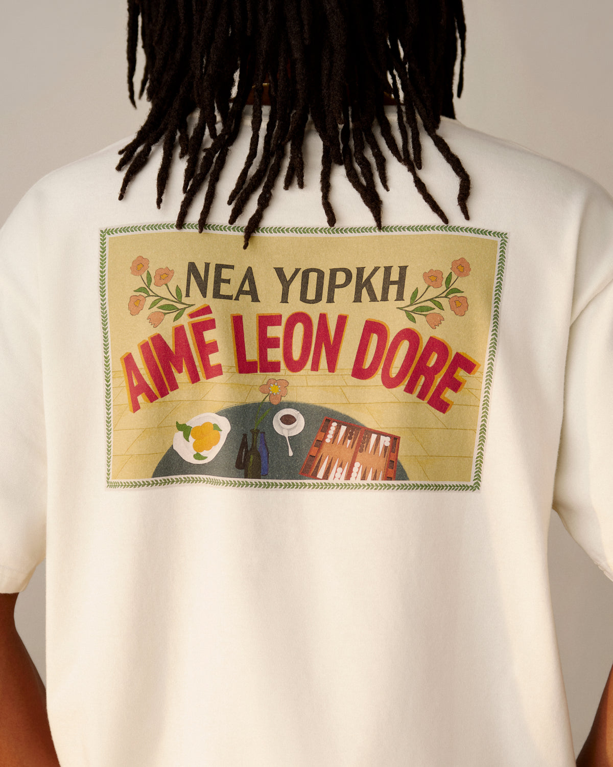 Person wearing a white Aimé Postcard Tee with food and building graphics on the back.