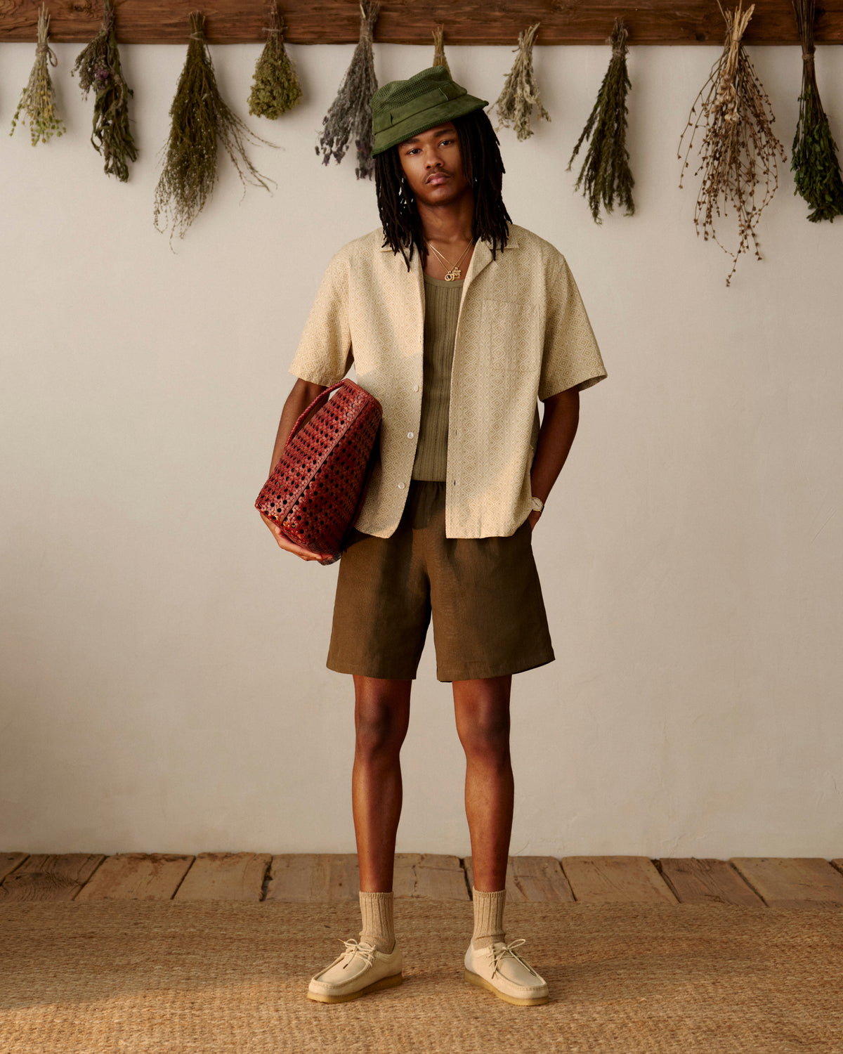 Wearing Linen Leisure Shorts, a person stands indoors by dried herbs, holding a red woven bag.