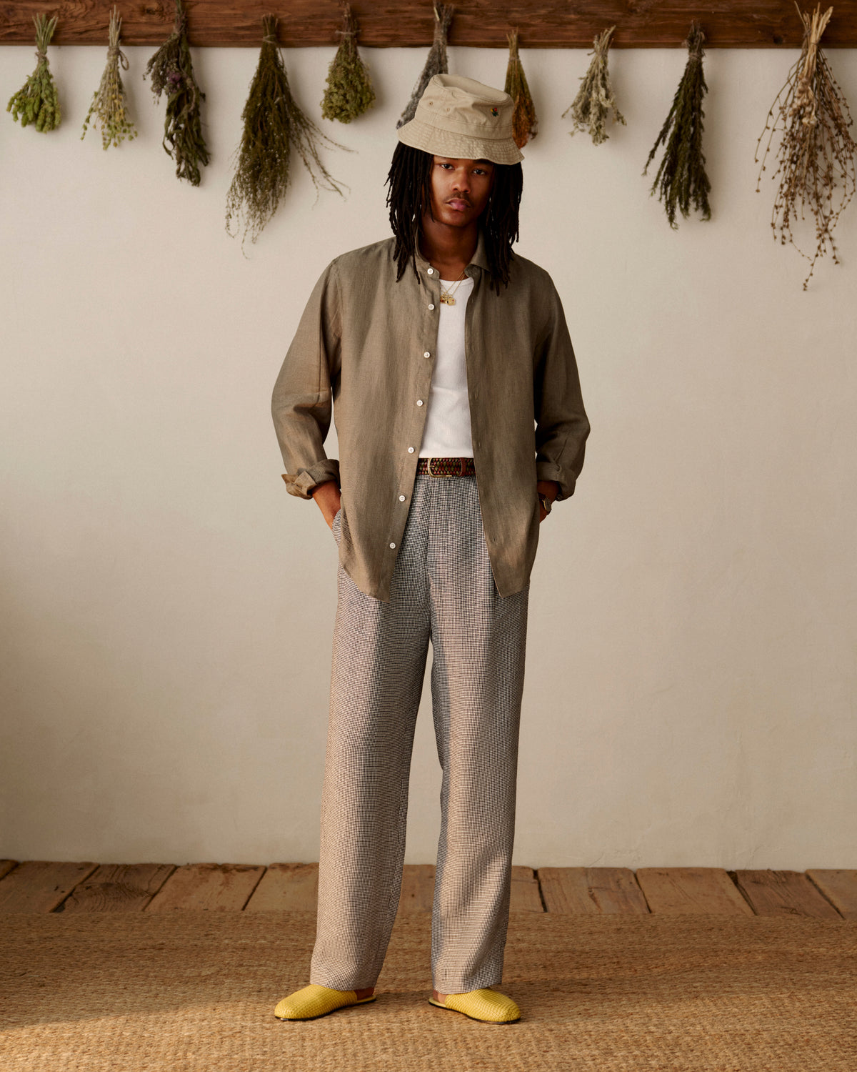 Wearing Houndstooth Pleated Trousers, a person stands indoors with dried herbs hanging on the wall behind.