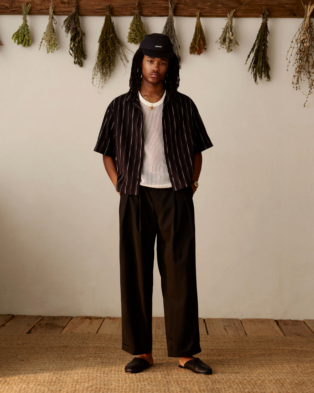 Man wears Mesh V-Neck Tee, wide black pants, and hat indoors with herbs hanging on the wall behind him.