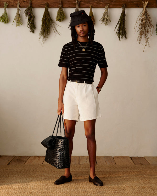A person in a black hat, striped shirt, white shorts, and loafers stands with a Woven Leather Tote Bag in a rustic room.