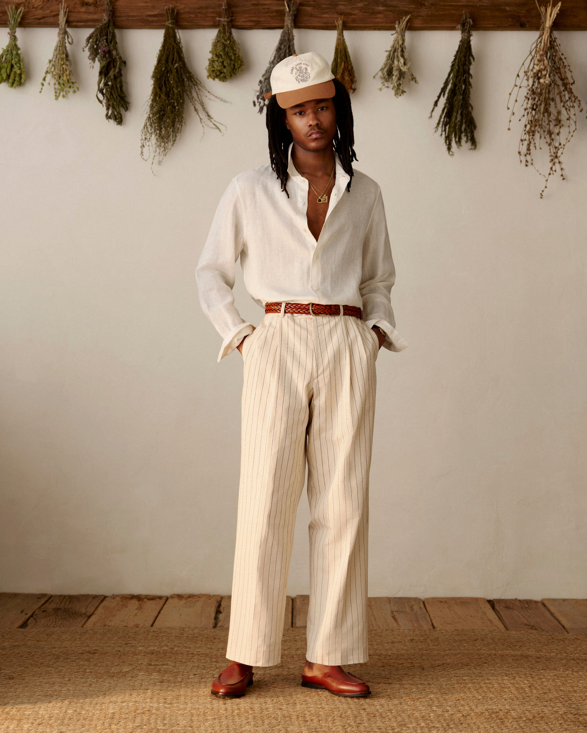 A person wears Double Pleated Pinstripe Suit Trousers, a white shirt, cap, and loafers, standing indoors by dried herbs.