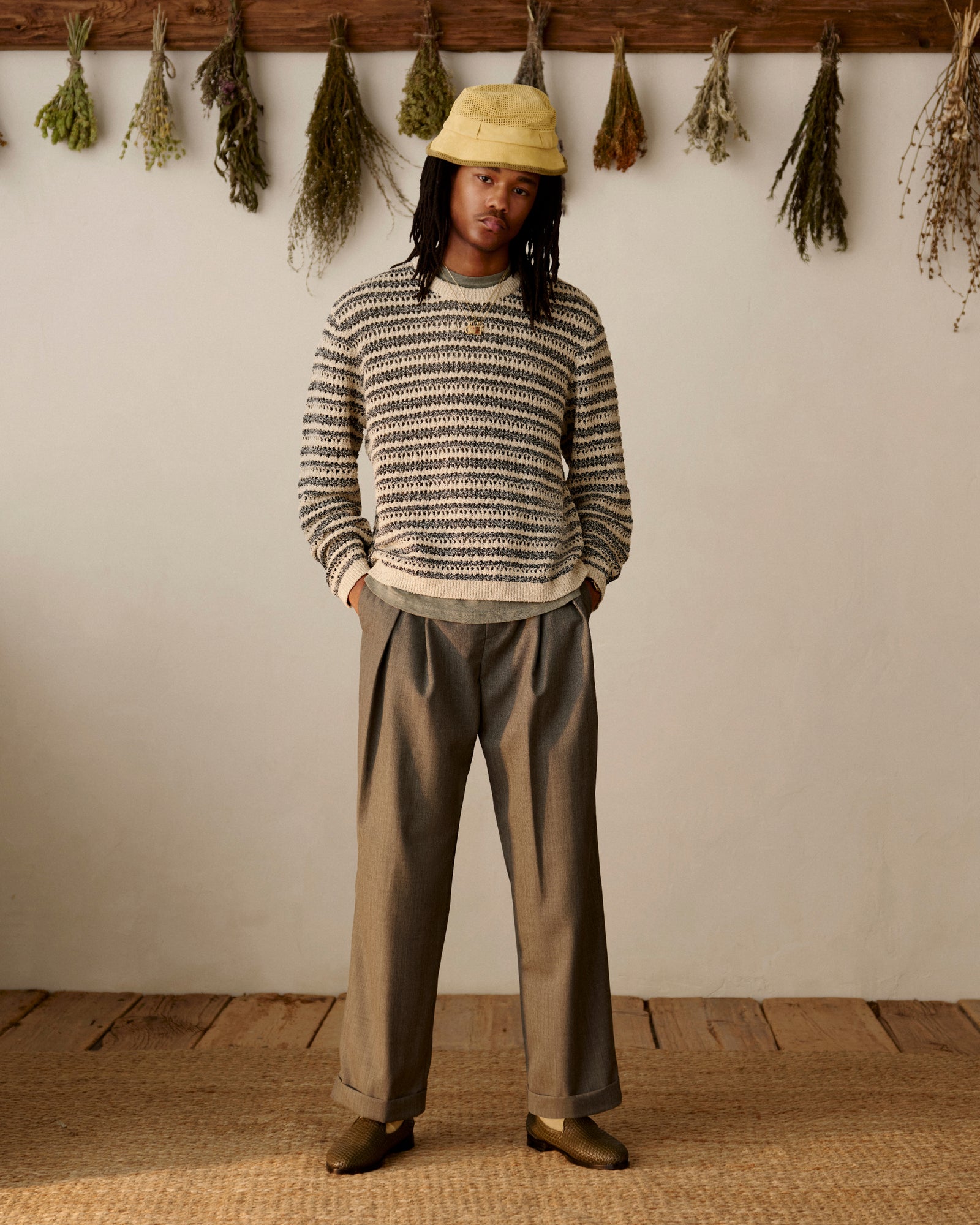 Person in a Bouclé Open Knit Crewneck Sweater, loose trousers, and loafers stands indoors near dried herbs on the wall.