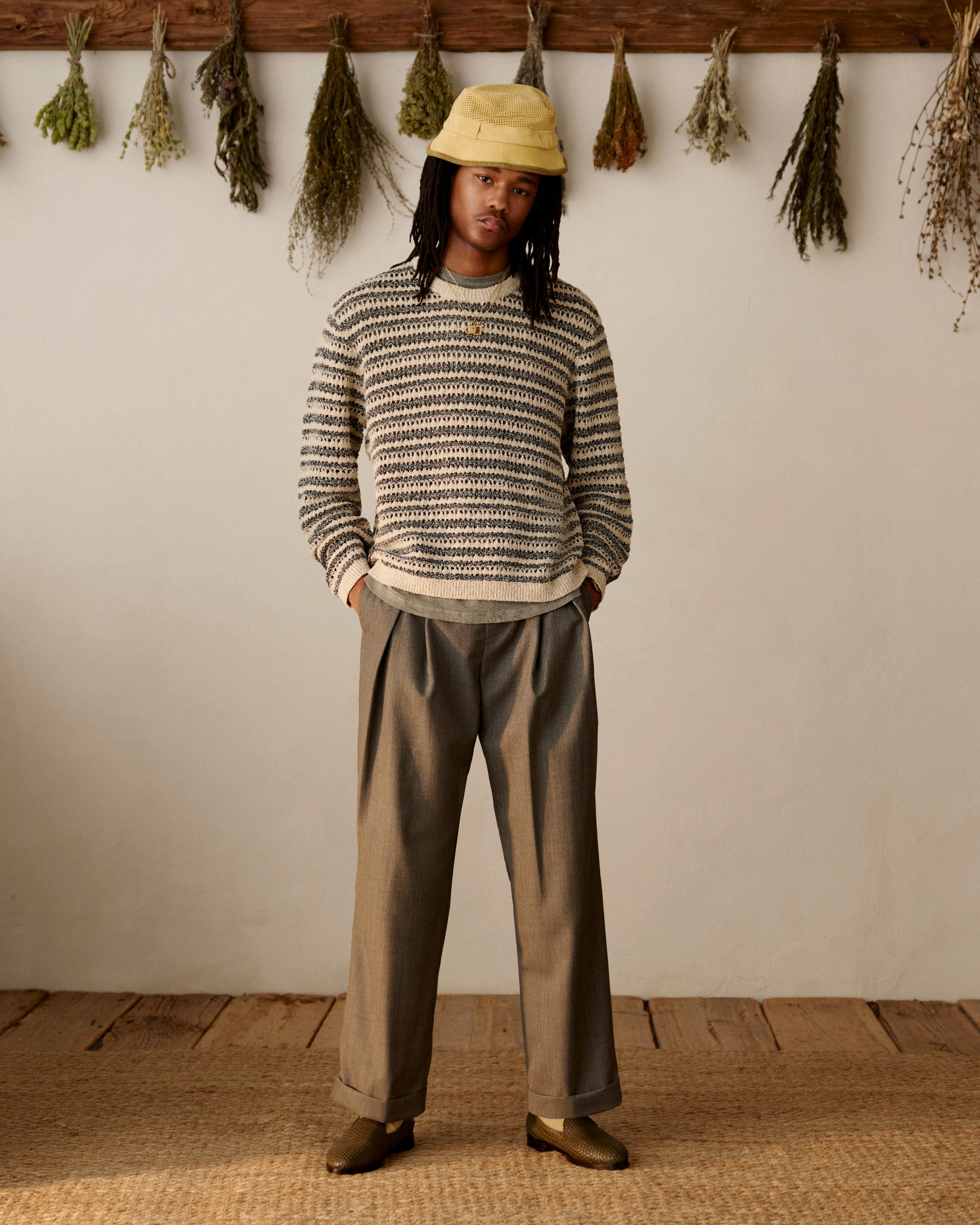Person in a Bouclé Open Knit Crewneck Sweater, loose trousers, and loafers stands indoors near dried herbs on the wall.