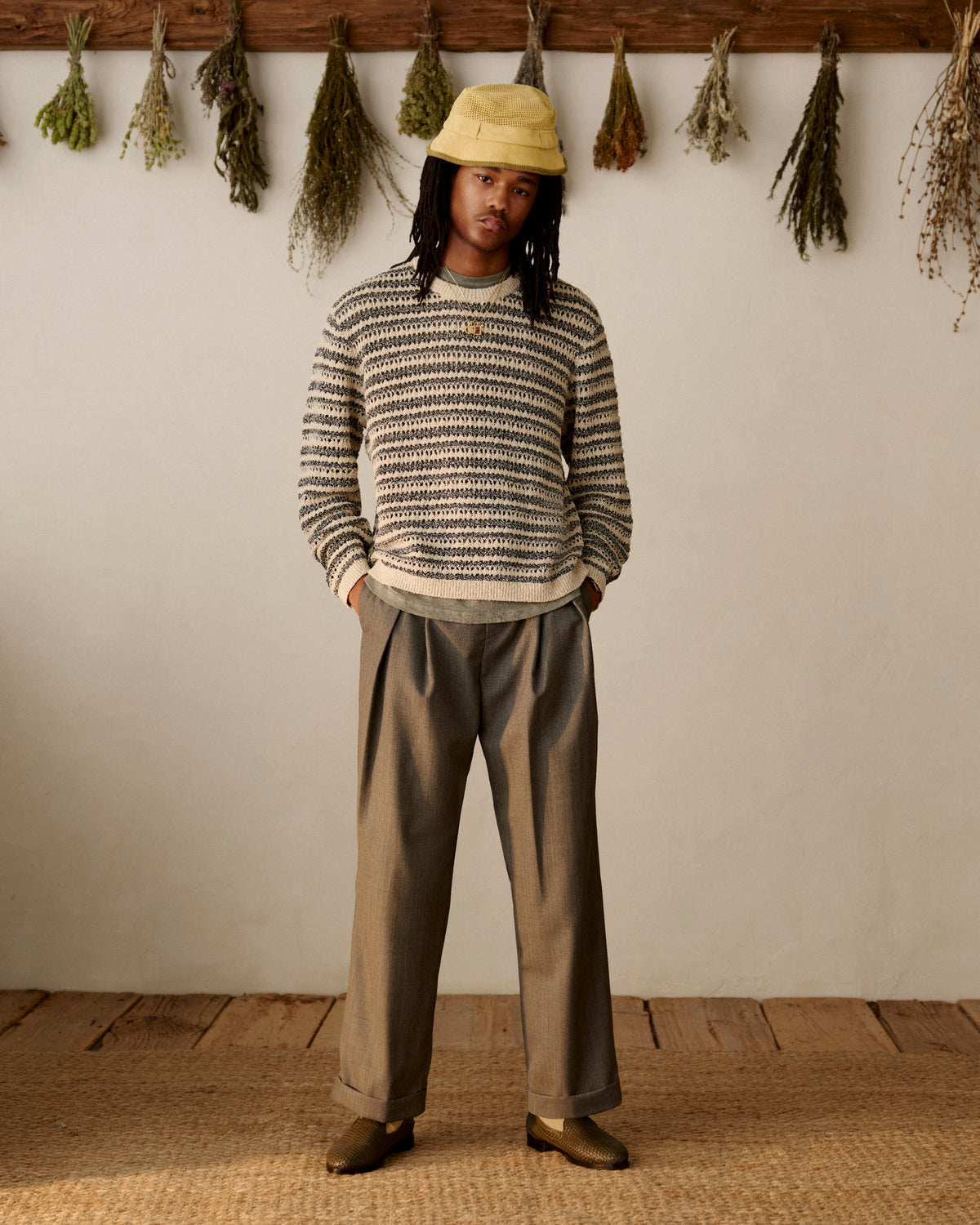 Person in striped sweater, yellow bucket hat, loafers, and Lightweight Cuffed Trouser stands indoors by hanging dried herbs.