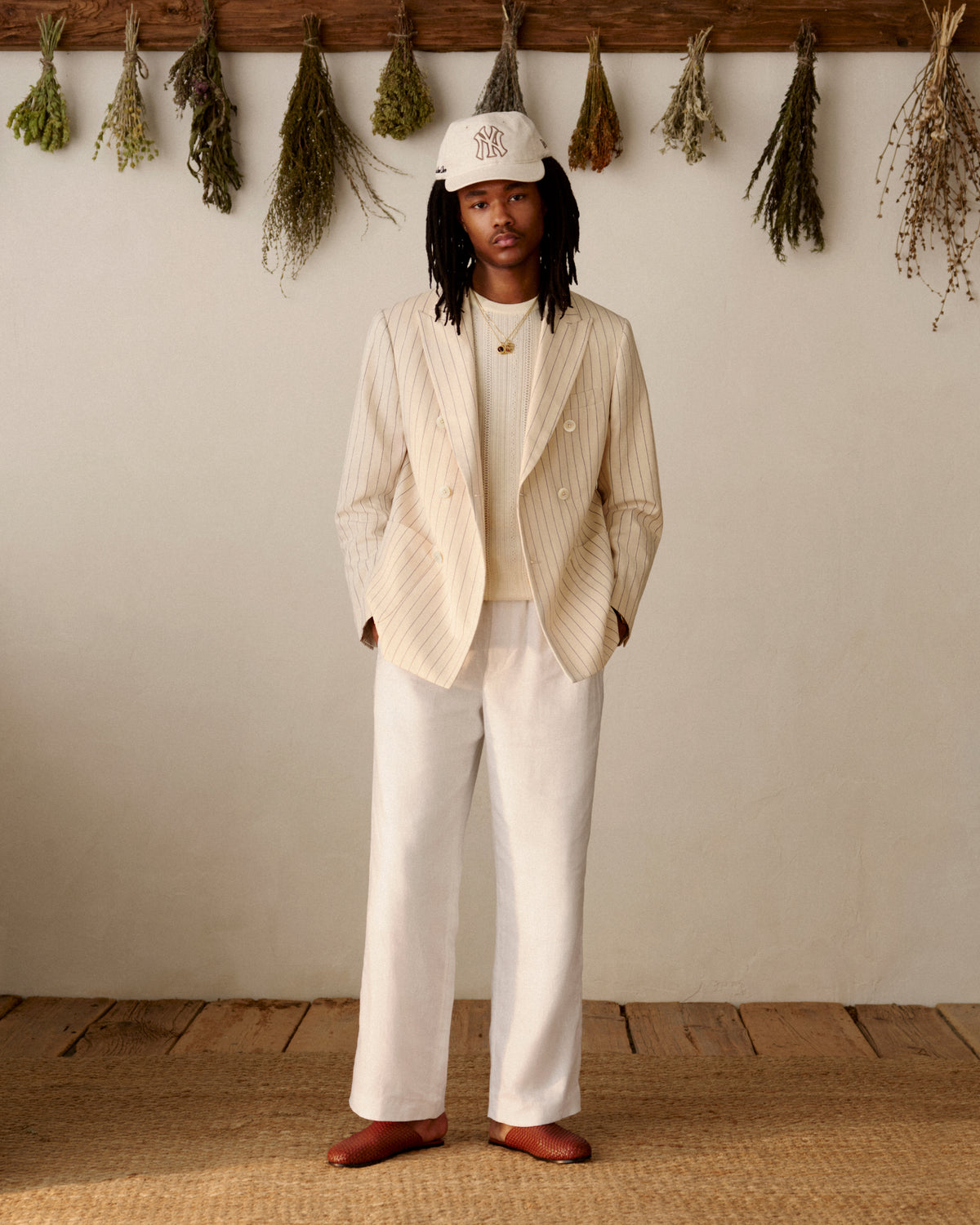 A man in a cream pinstripe suit and white shirt wears Leather Raffia Mules, standing indoors by dried herbs on the wall.
