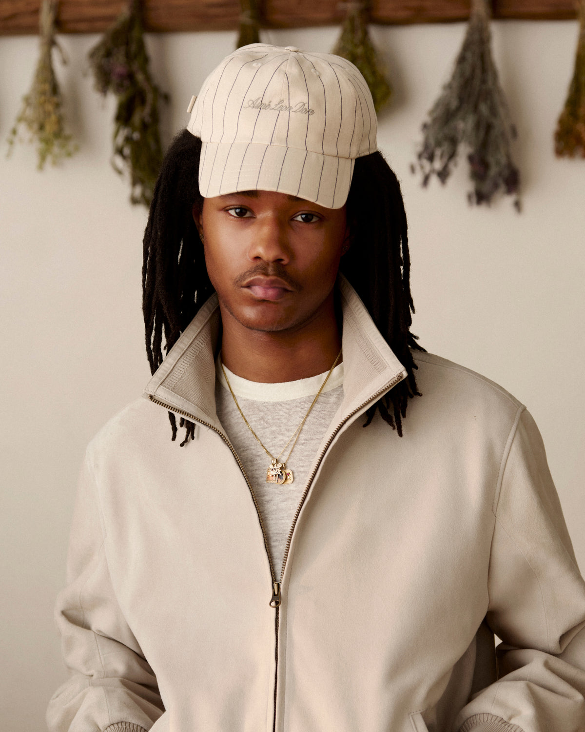 A man in a beige jacket and a Pinstripe Suiting Hat stands indoors with dried herbs hanging behind him.