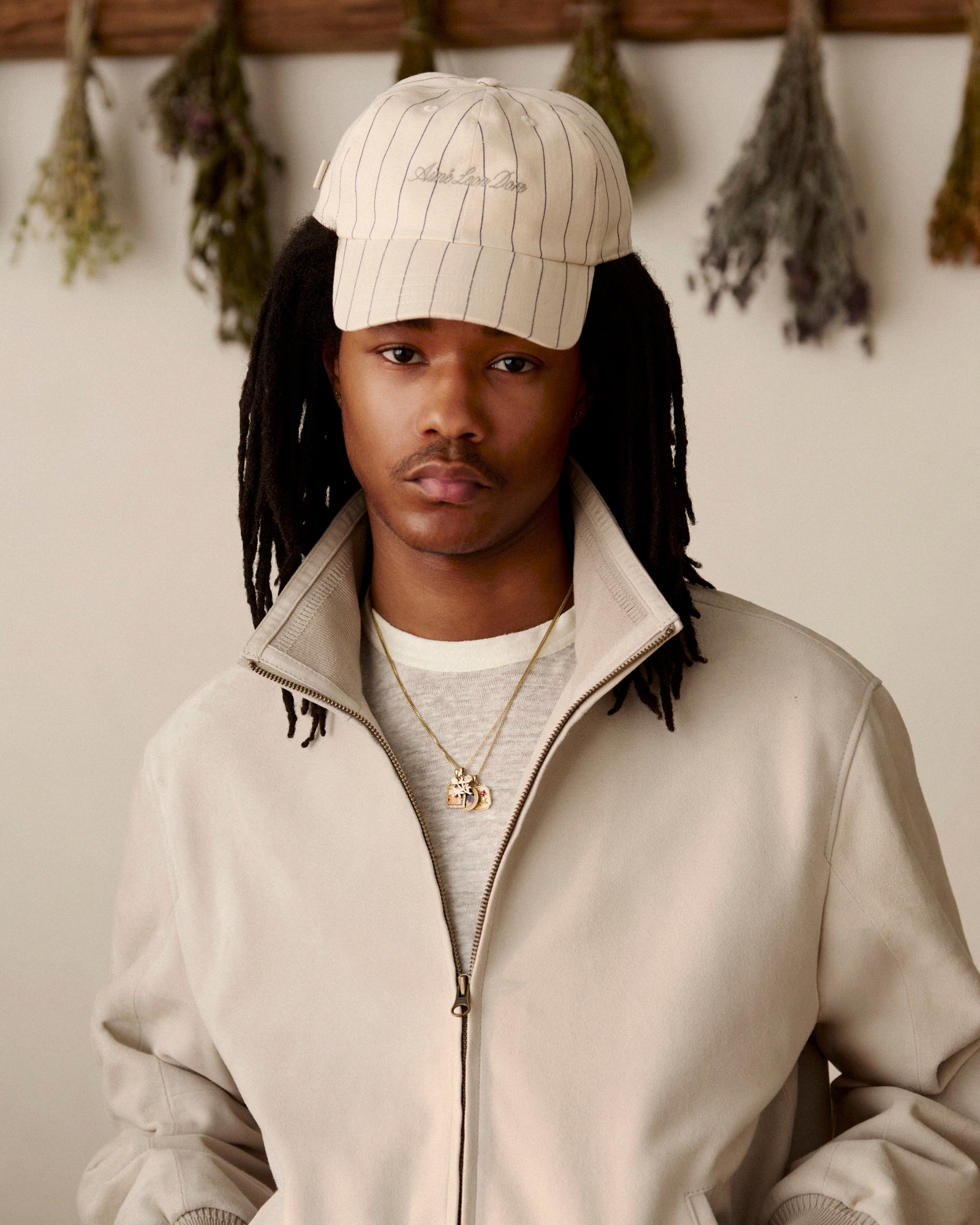 A man in a beige jacket and a Pinstripe Suiting Hat stands indoors with dried herbs hanging behind him.