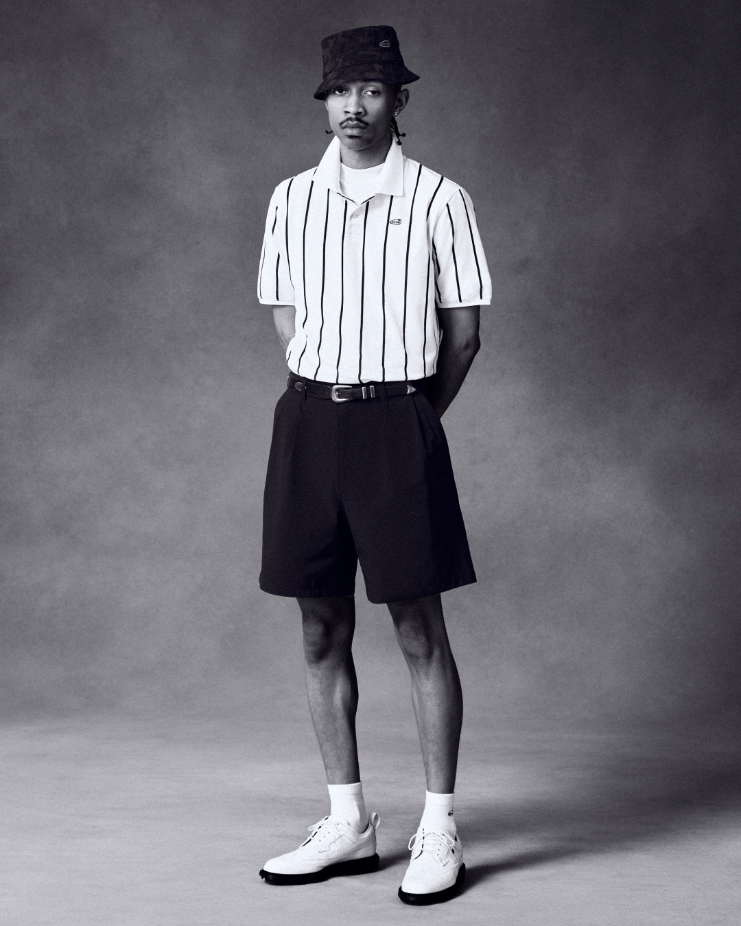 Man wears ALD Golf Polo, black shorts, bucket hat, and white shoes while standing against a plain backdrop.