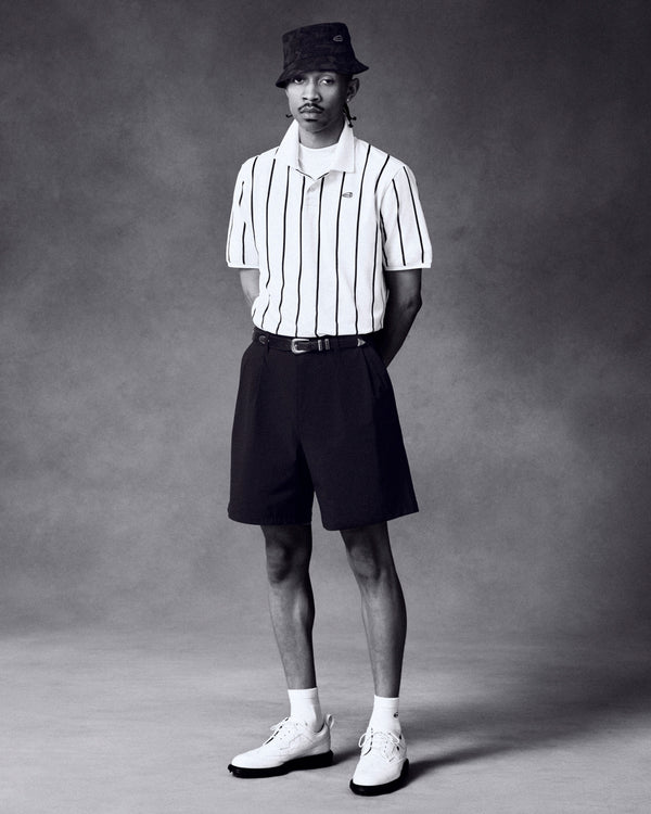 Man in striped polo, ALD Golf Cordura Double Pleated Short, bucket hat, and white shoes against plain studio background.