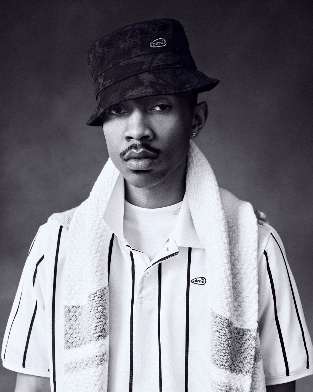 Black-and-white portrait of a man in an ALD Golf Bucket Hat, striped polo, and towel draped over his shoulders, looking serious.