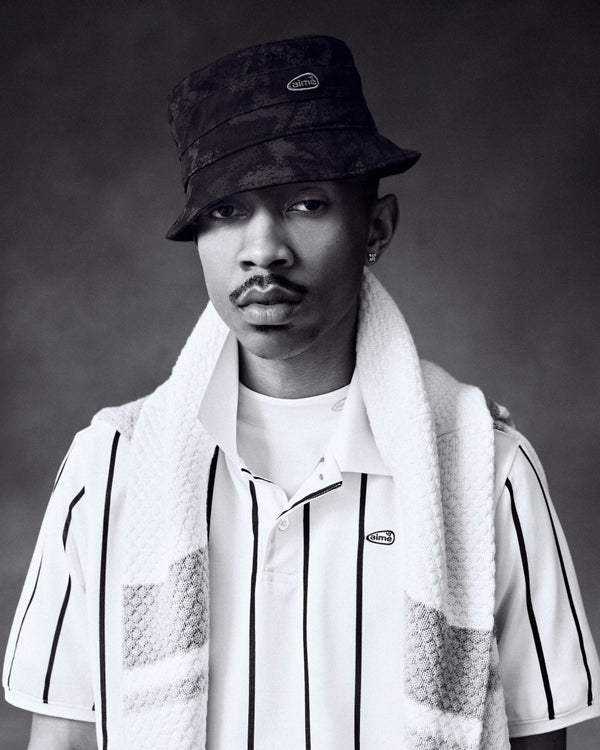 Black-and-white portrait of a man in an ALD Golf Bucket Hat, striped polo, and towel draped over his shoulders, looking serious.