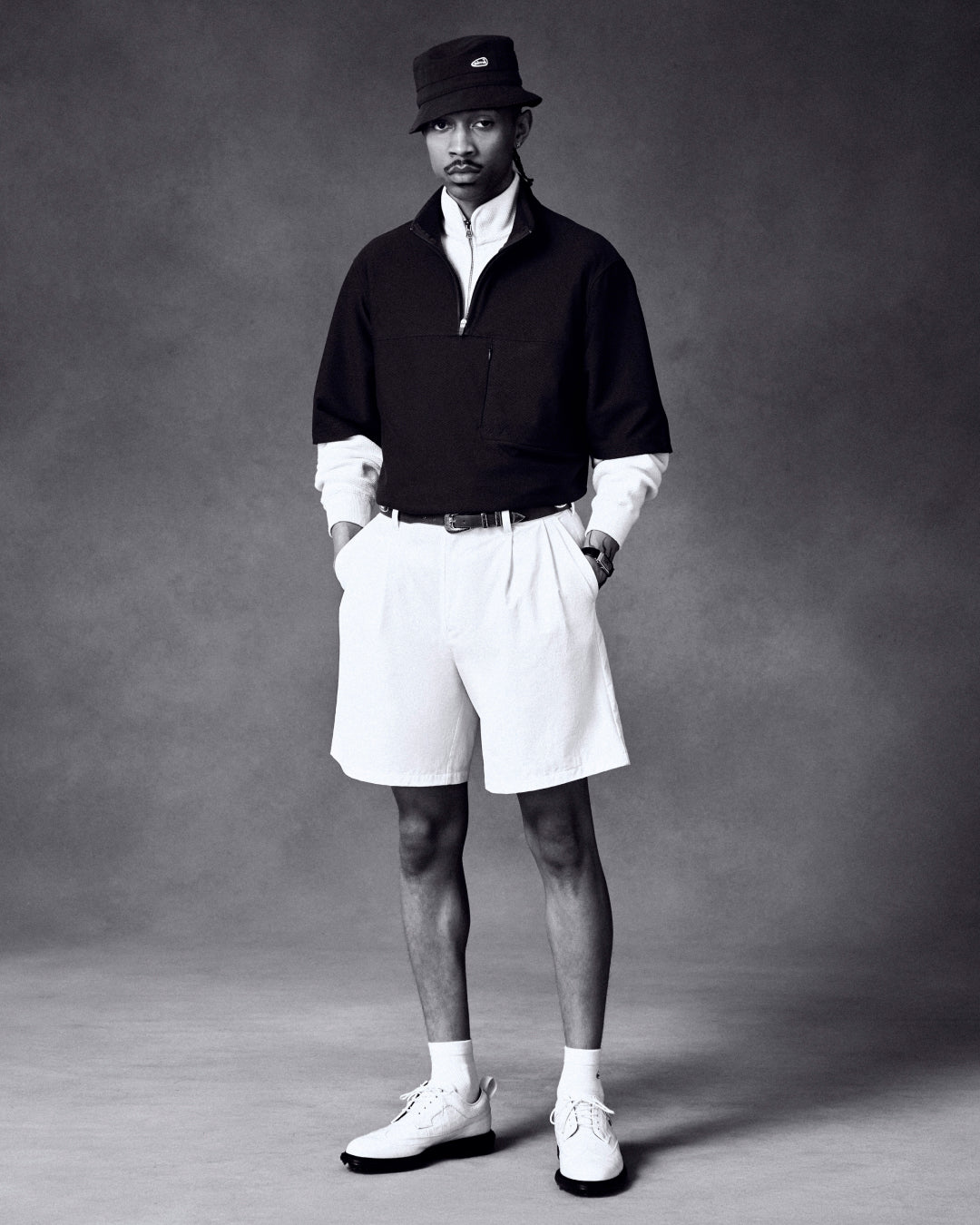A man stands in a studio wearing an ALD Golf Bucket Hat, dark pullover, white shirt, shorts, and shoes.