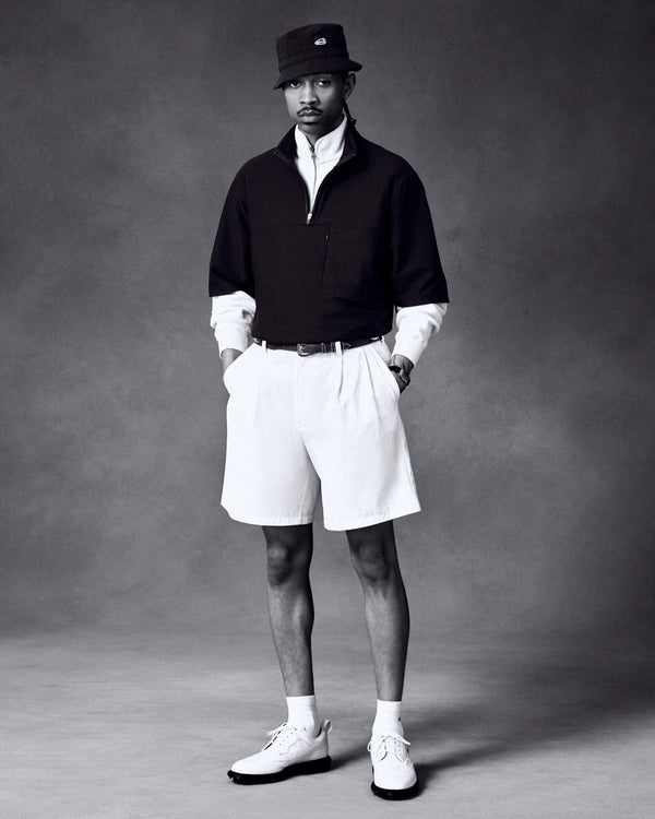 A man stands in a studio wearing an ALD Golf Bucket Hat, dark pullover, white shirt, shorts, and shoes.