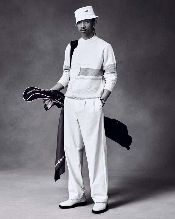 Black-and-white photo of a man in a bucket hat holding golf clubs, wearing the ALD Golf Clubhouse Sweater.