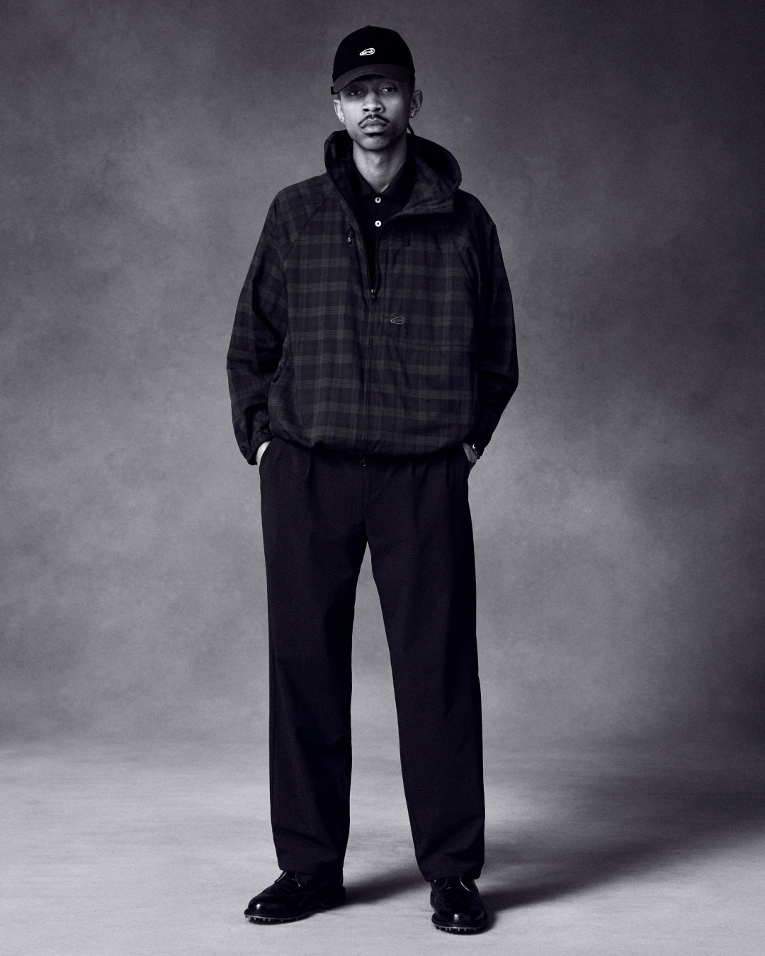 A man wearing the ALD Golf Lightweight Tips Windbreaker, black pants, shoes, and cap stands with hands in pockets.