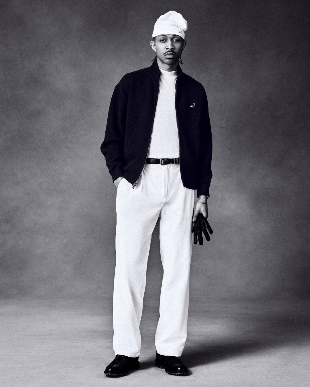 A man in a white hat, white shirt, pants, and dark jacket holds an ALD / FootJoy StaSof Heritage Glove (Left).