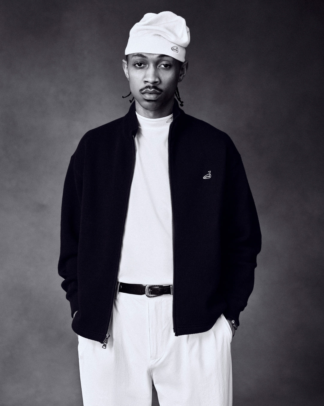 A man in an ALD Golf Skull Cap, white shirt, black jacket, and white pants stands with hands in pockets.