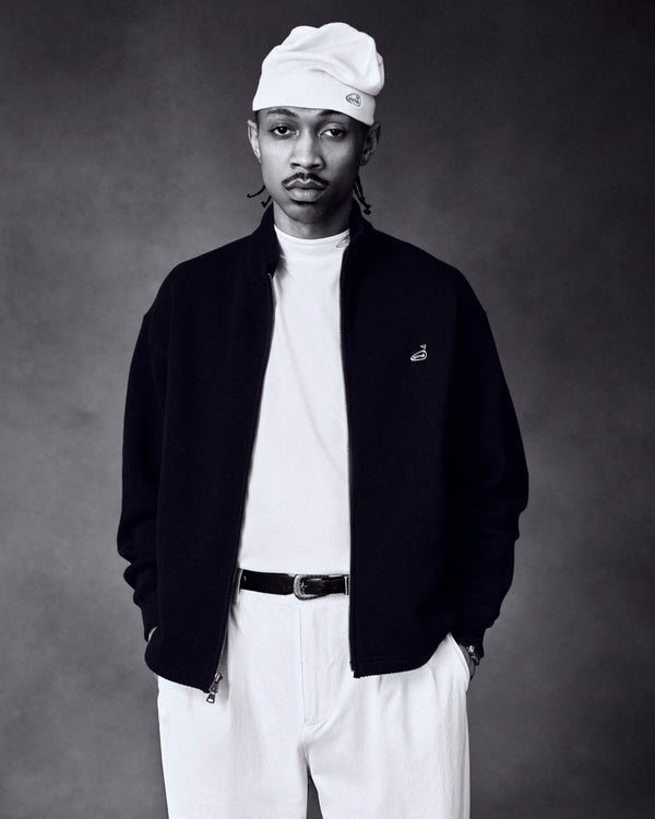 Man in ALD Golf Performance Tee, light beanie, dark jacket, and white pants, hands in pockets against dark background.