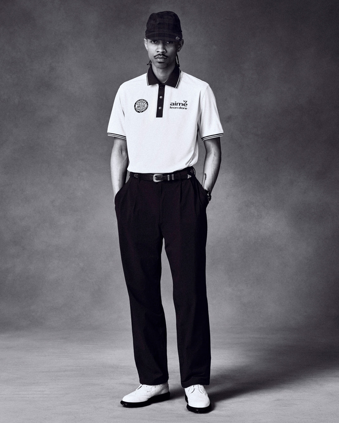 A man in a white polo, dark pants, ALD Golf Performance Sport Hat, and white shoes stands against a gray backdrop.