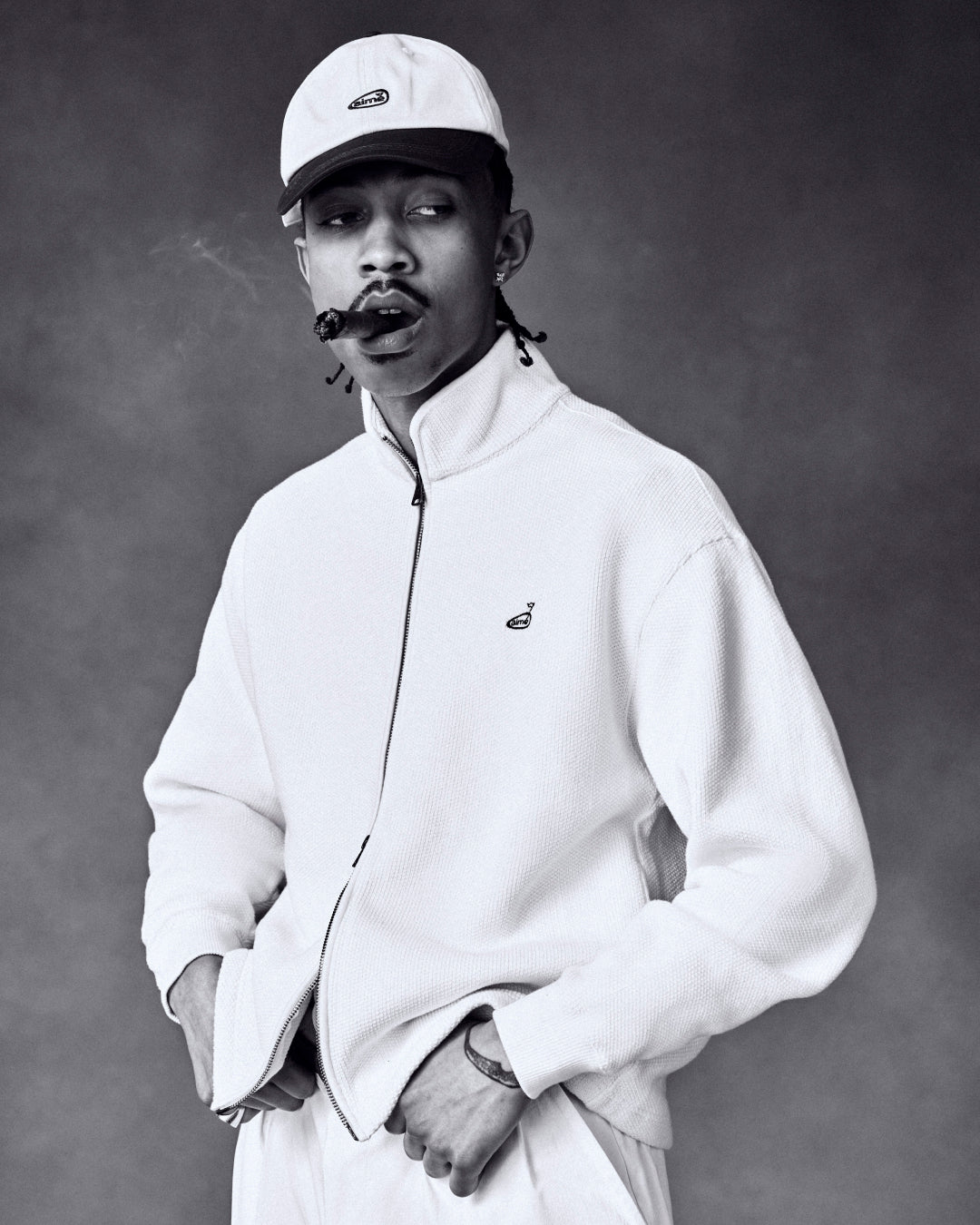 Man wearing ALD Golf Logo Hat and light jacket smokes a cigar, looking sideways against a plain studio backdrop.