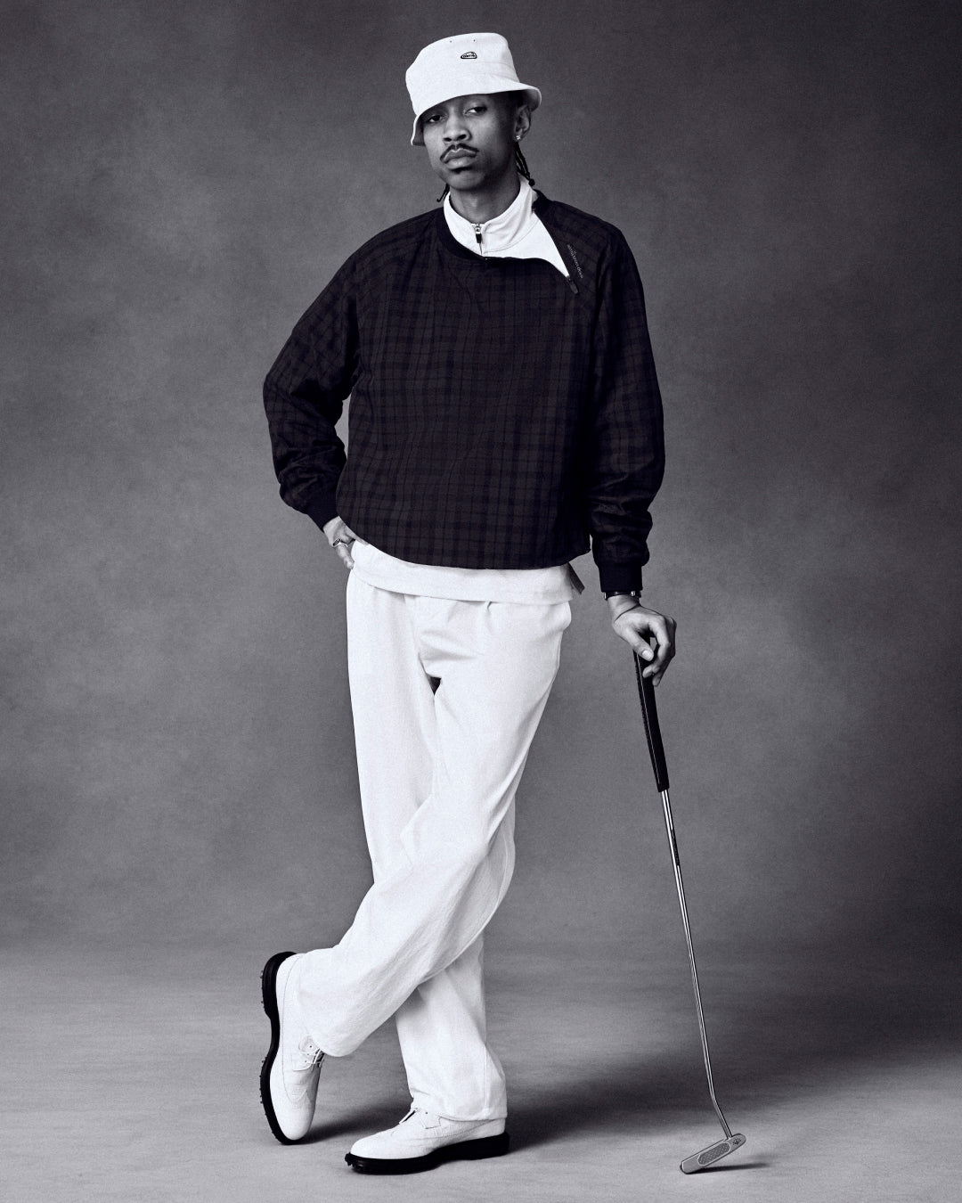 Man wears ALD Golf Cordura Double Pleated Pant, dark sweater, and bucket hat while posing with golf club in studio.