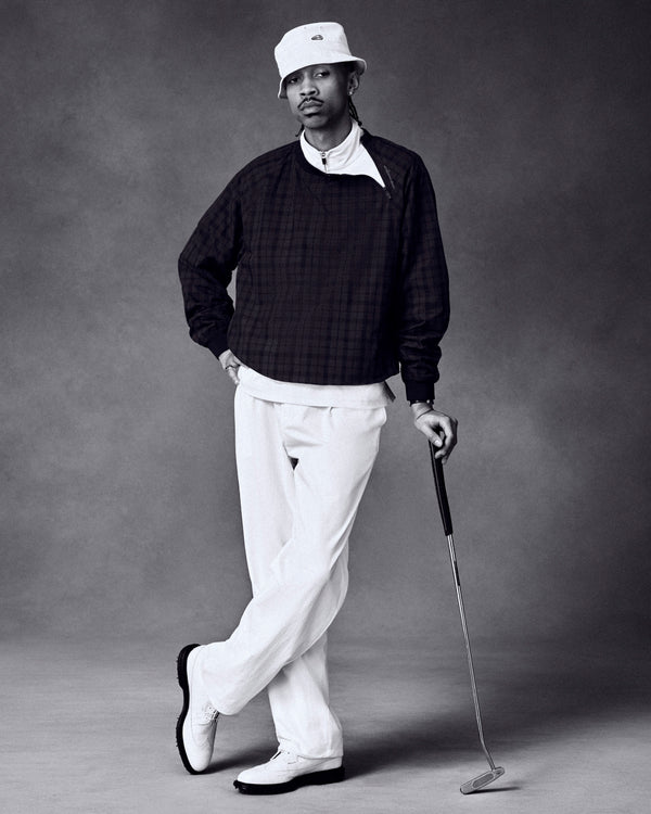 Man wears ALD Golf Cordura Double Pleated Pant, dark sweater, and bucket hat while posing with golf club in studio.