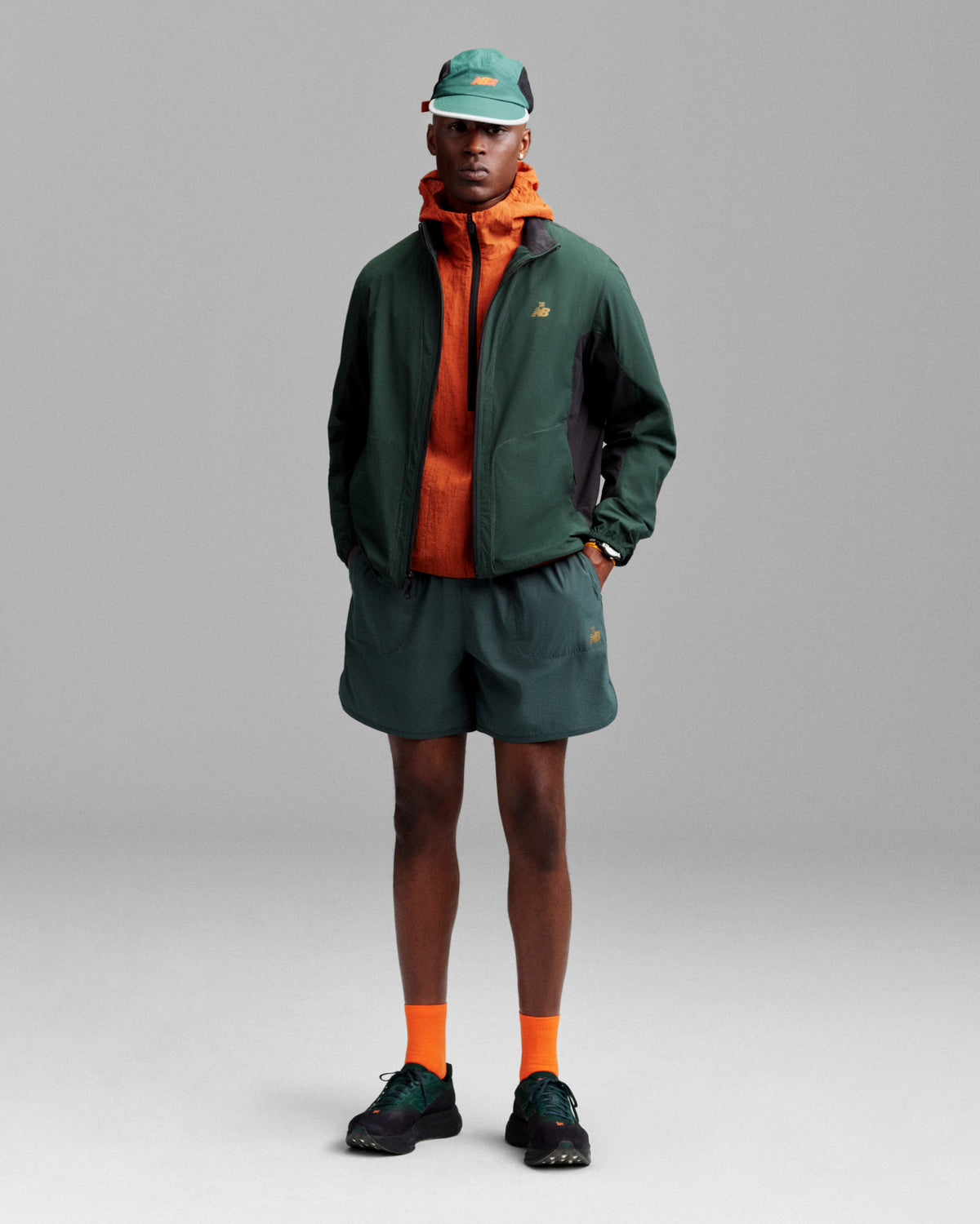 A person in a green jacket, orange hoodie, green shorts, orange socks, black shoes, and an ALD/New Balance Mesh Sport Hat stands facing forward.