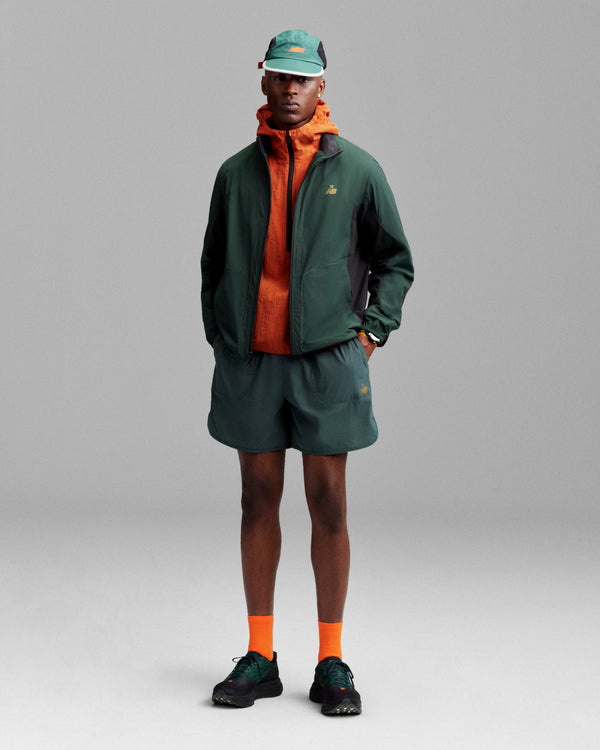 A person in a green jacket, orange hoodie, green shorts, orange socks, black shoes, and an ALD/New Balance Mesh Sport Hat stands facing forward.