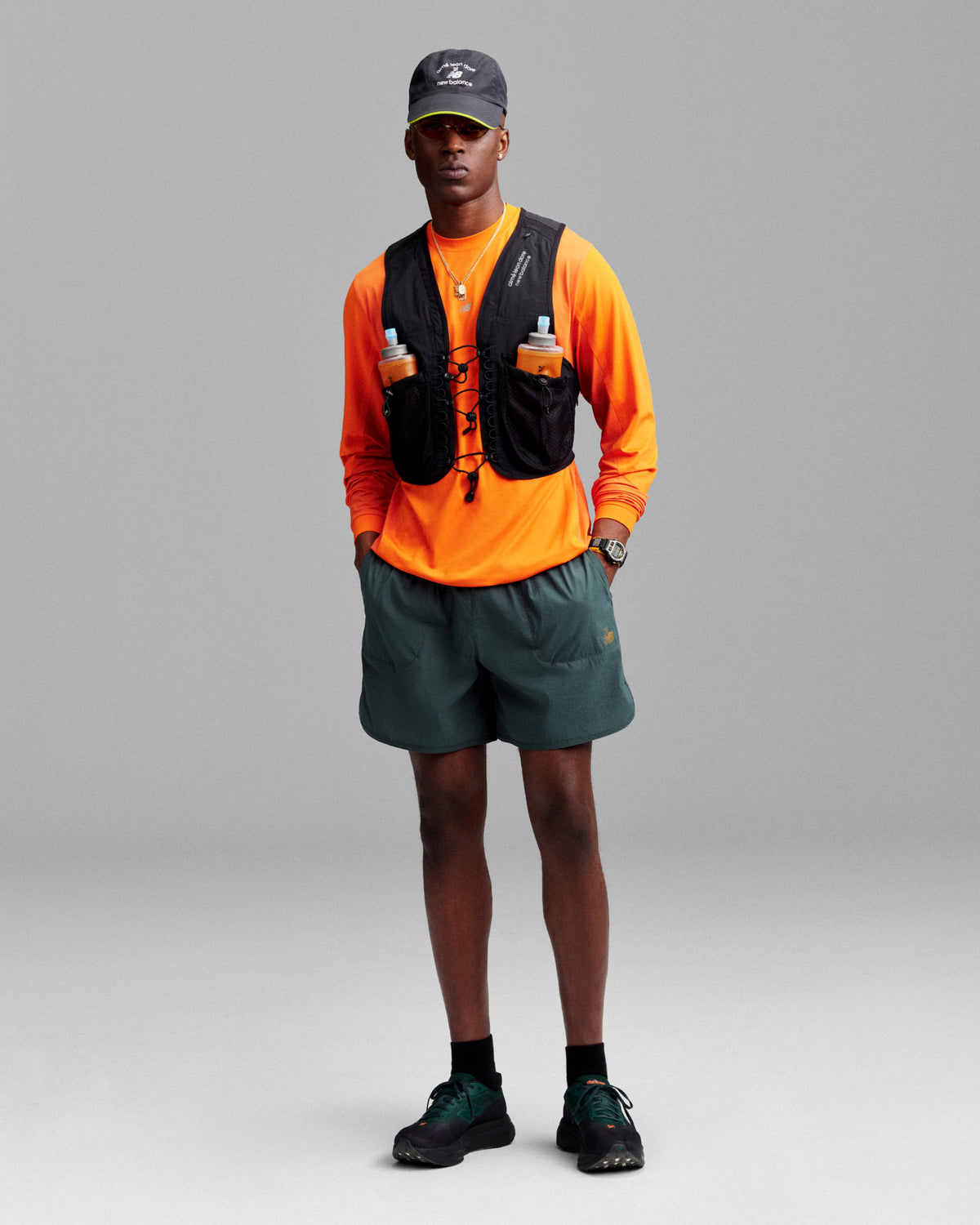 A person in an ALD / New Balance Performance Long-Sleeve Tee: orange shirt, green shorts, black vest, hat, sneakers.