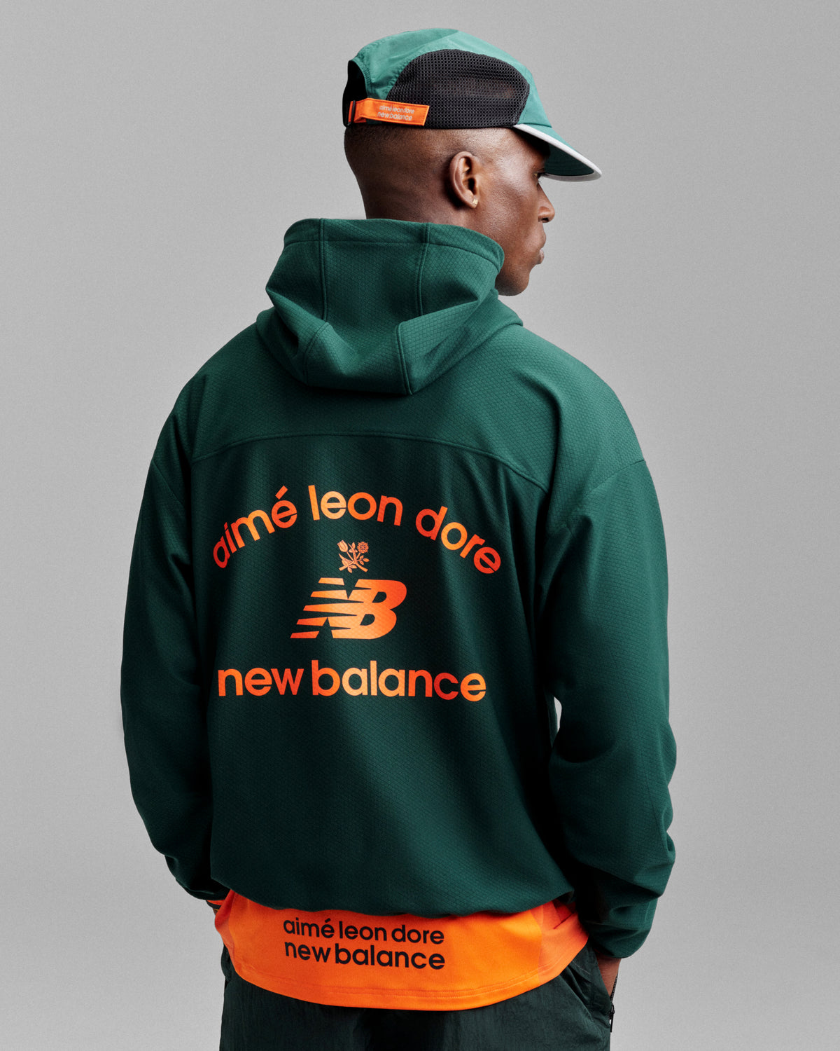 Man in green ALD / New Balance Full-Zip Warmup Hoodie and cap, orange logo on back, facing away.