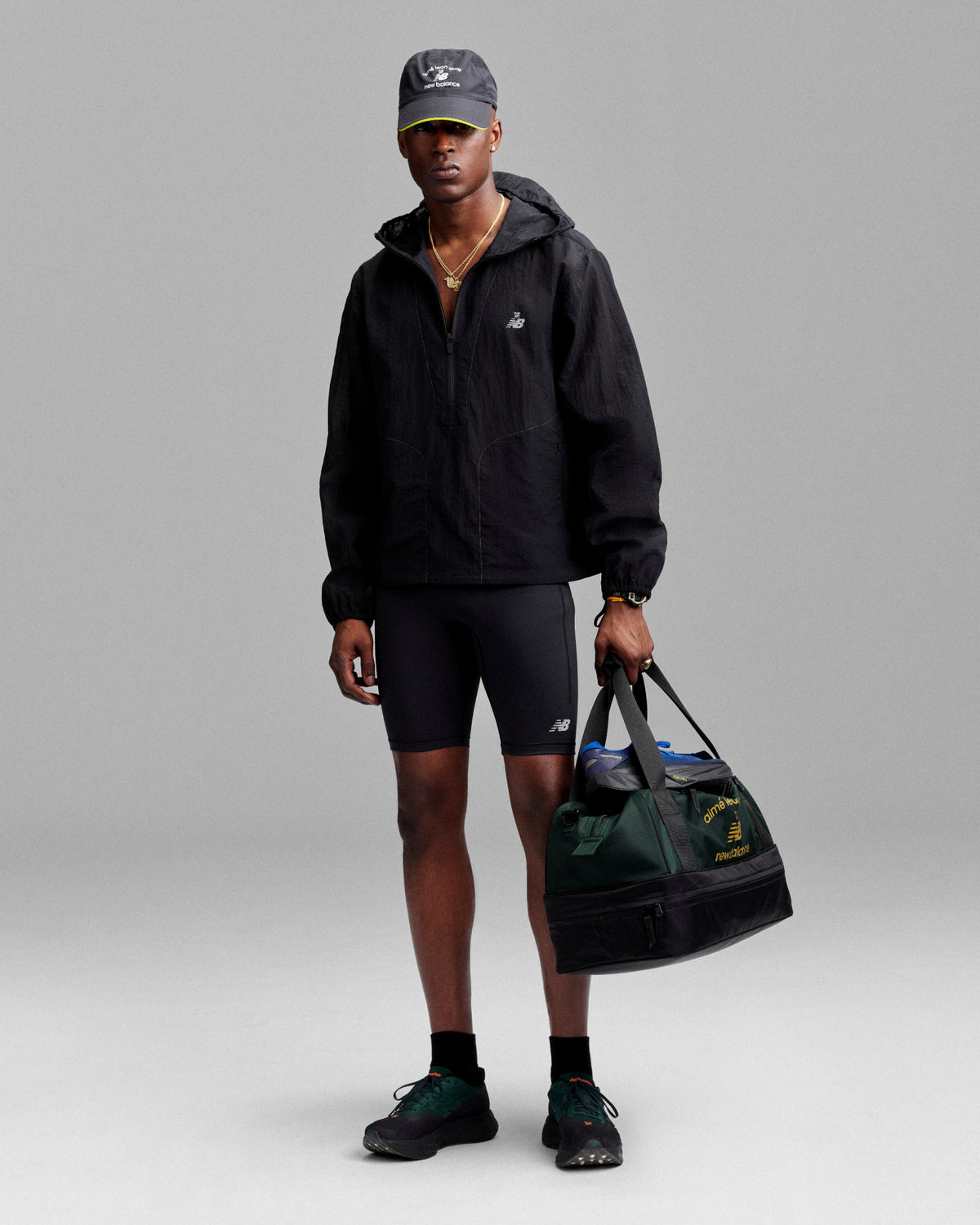 Man in black jacket, bike shorts, and cap holds an ALD / New Balance Travel Duffle Bag against a gray background.