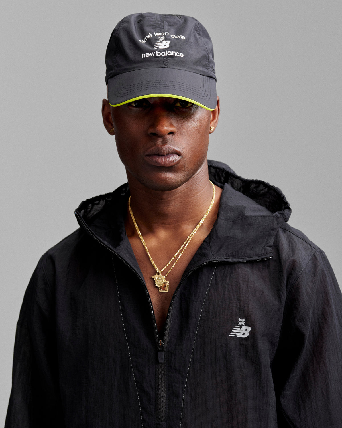 A man in a black jacket and ALD / New Balance Running Hat, with gold necklaces, poses against a gray background.