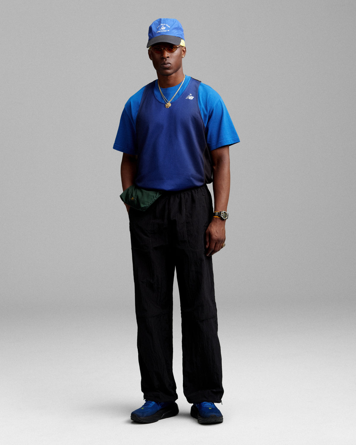 A man in an ALD / New Balance Running Bib Tee, blue cap, black pants, and blue sneakers stands by a gray background.