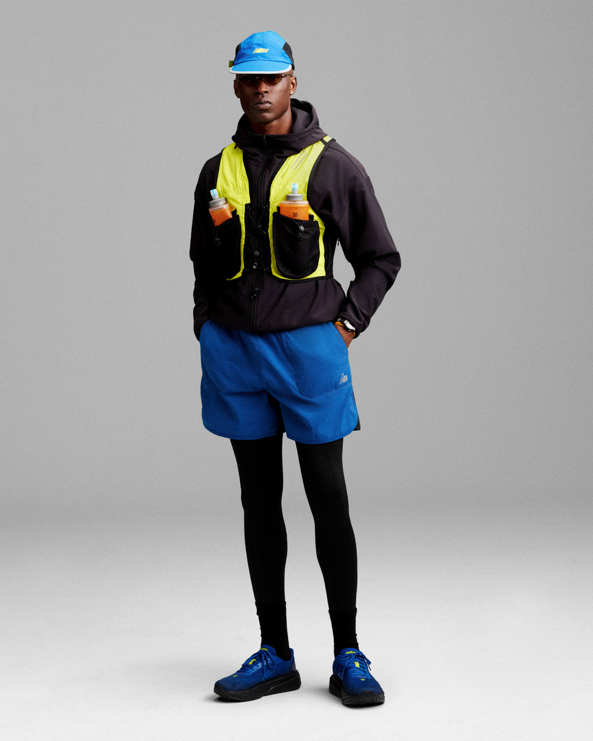 A person in running gear with an ALD / New Balance Full-Zip Warmup Hoodie, yellow vest, blue shorts, and hydration bottles.