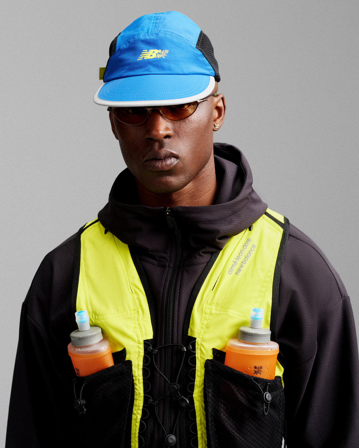 Someone wearing an ALD / New Balance Mesh Sport Hat, sunglasses, black jacket, yellow vest, holding orange bottles.