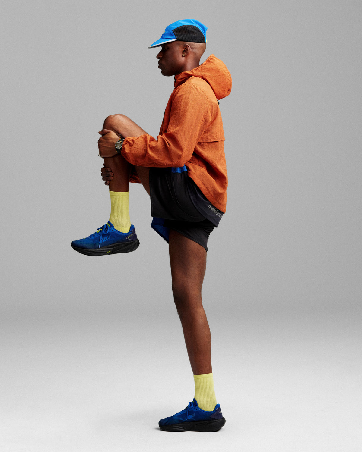 Man in orange jacket, blue cap, yellow socks, and ALD / New Balance FuelCell Rebel v5 stretching on gray background.