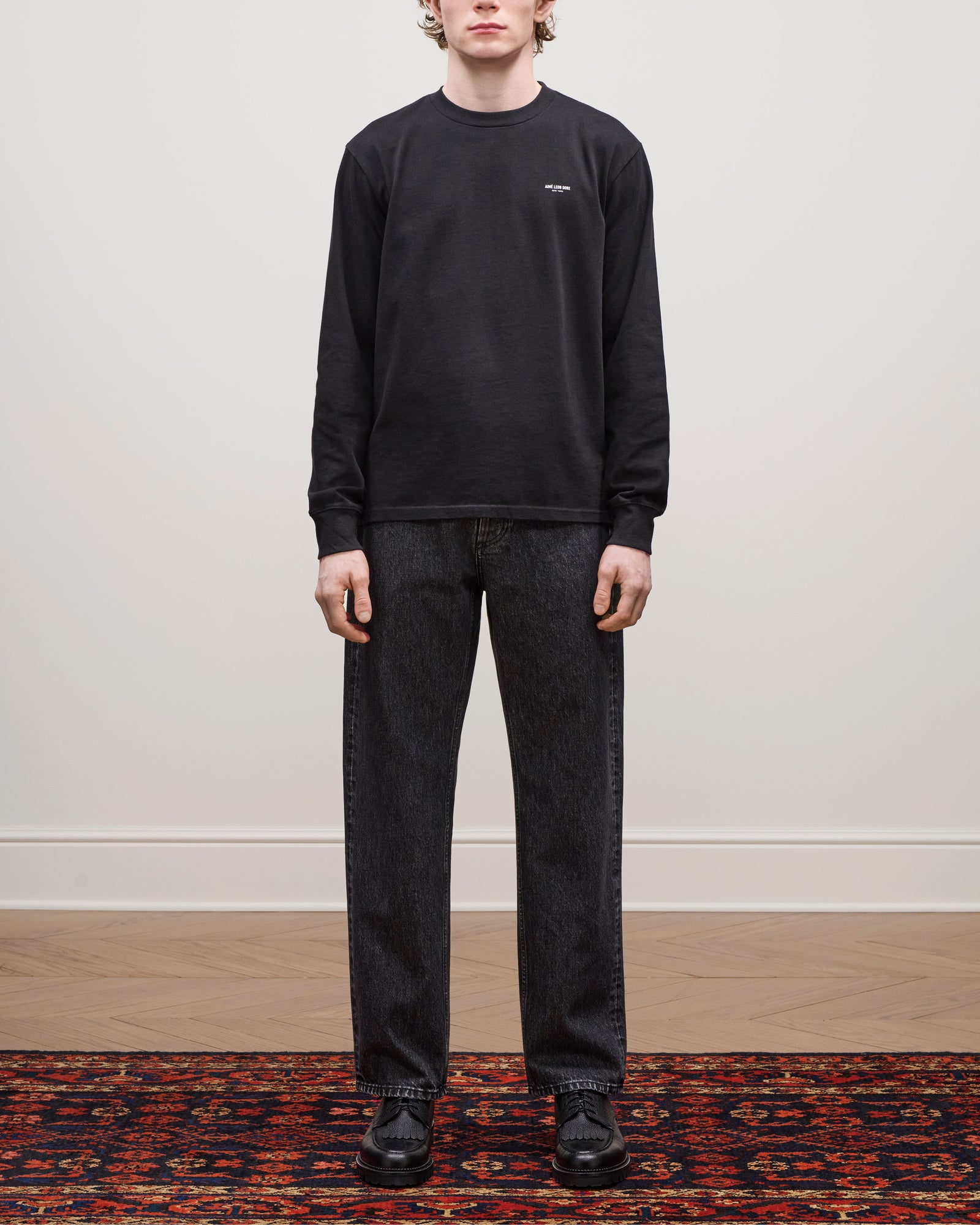 Person standing on a patterned rug wearing the Long-Sleeve Micro Logo Tee, black jeans, and black shoes.
