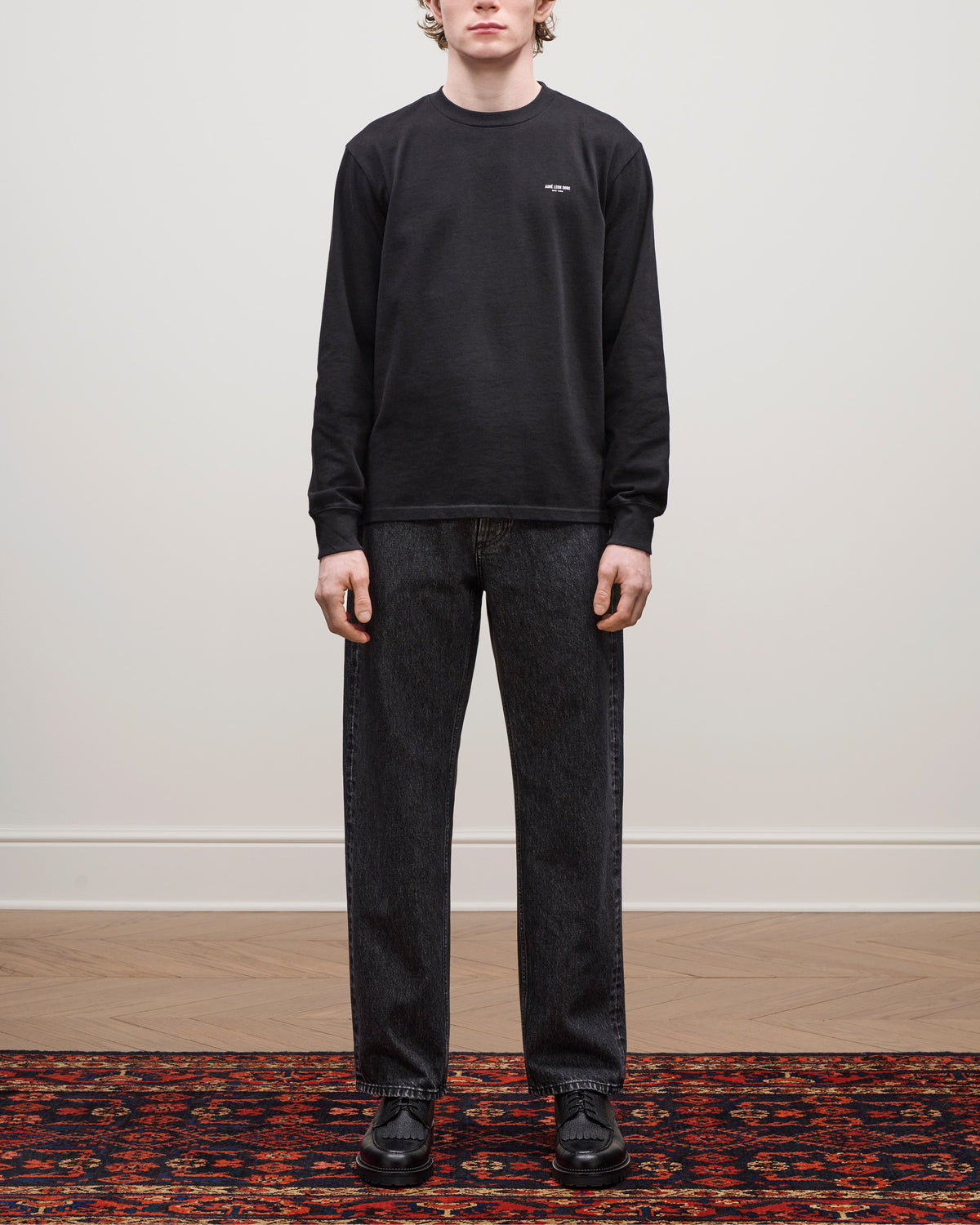 A person stands indoors on a patterned rug wearing Relaxed Fit 5-Pocket Denim Pant, a black sweatshirt, and black shoes.