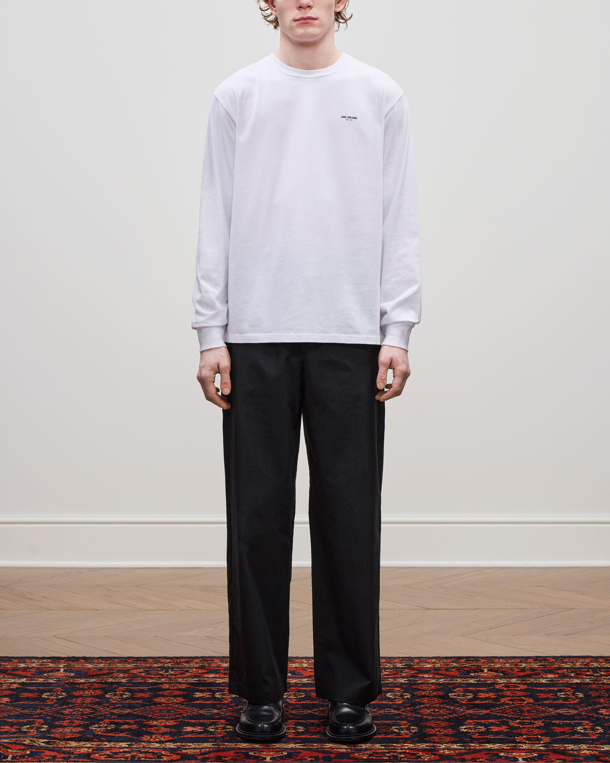 A person stands on a patterned rug wearing a white long-sleeve shirt and Elasticated Waist Gabardine Trouser, facing forward.