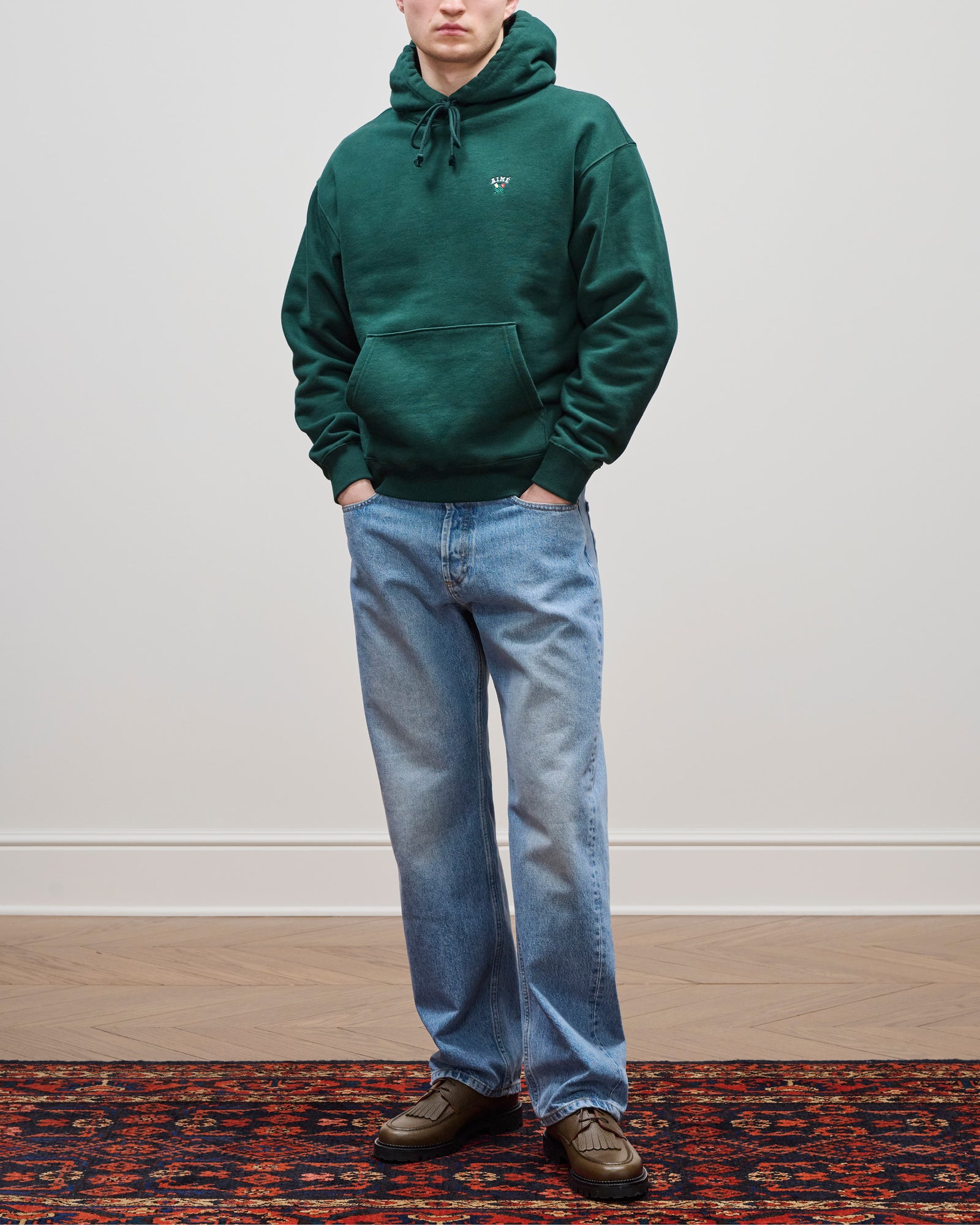 A person in a Crest Hoodie, light blue jeans, and brown shoes stands on a patterned rug against a plain wall.