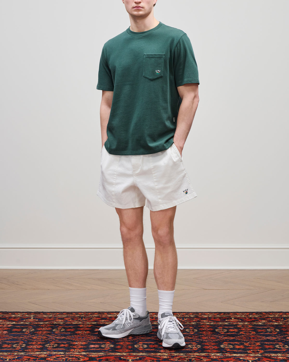 A person in a Crest Pocket Tee, white shorts, gray sneakers, and white socks stands on a patterned rug.