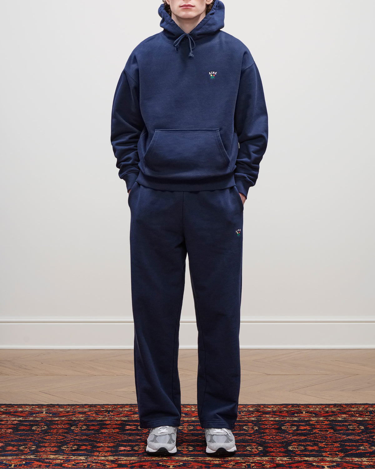 Person wears a Crest Hoodie in navy with matching sweatpants, standing on a patterned rug against a plain wall.