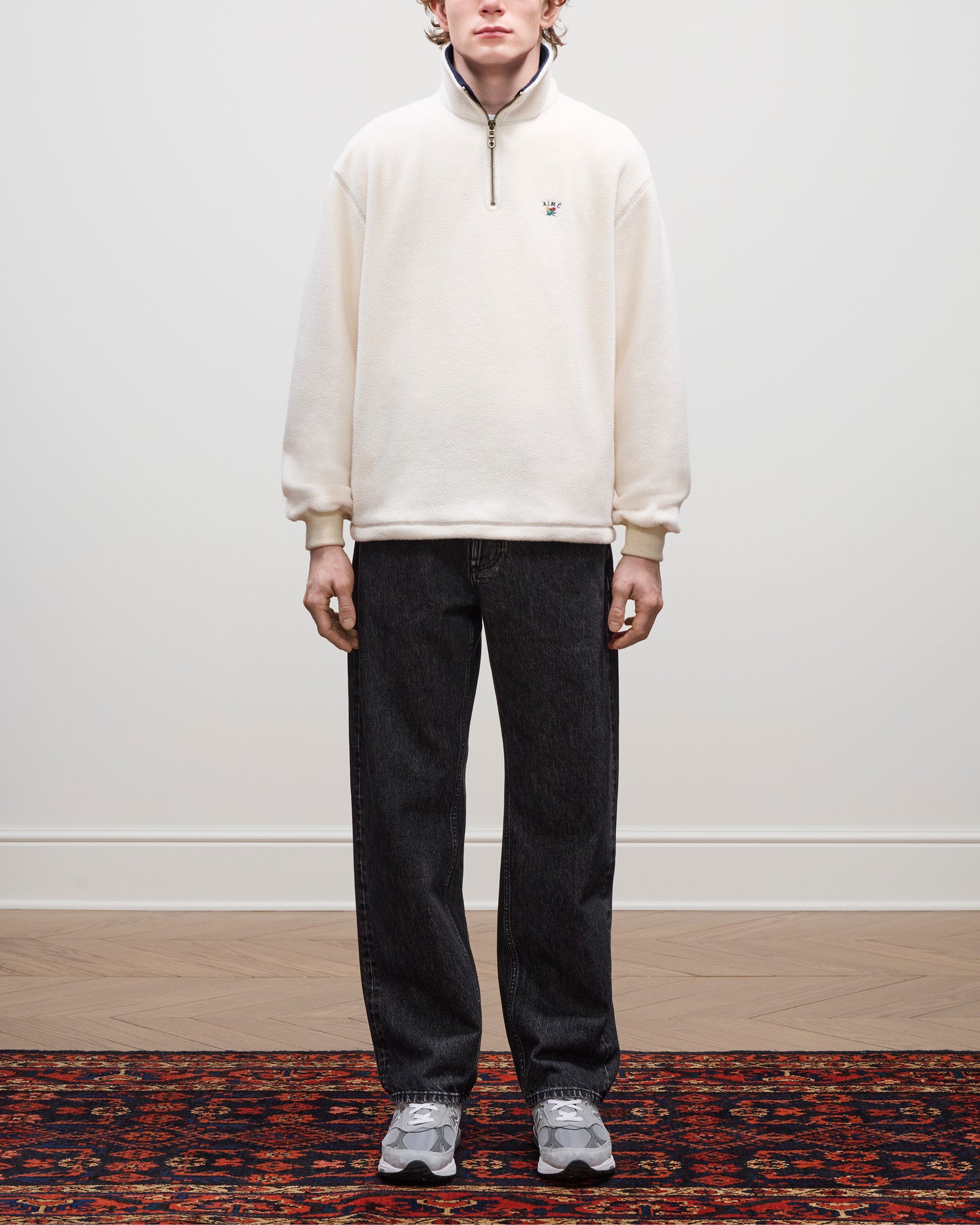 A person stands indoors wearing a Crest Quarter-Zip Fleece Pullover, dark jeans, and gray sneakers on a patterned rug.